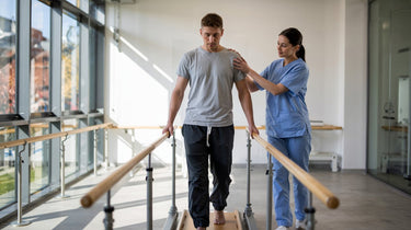 A male patient doing gait training in a modern rehabilitation center, walking barefoot between parallel bars with the support of a female physical therapist in medical scrubs. 